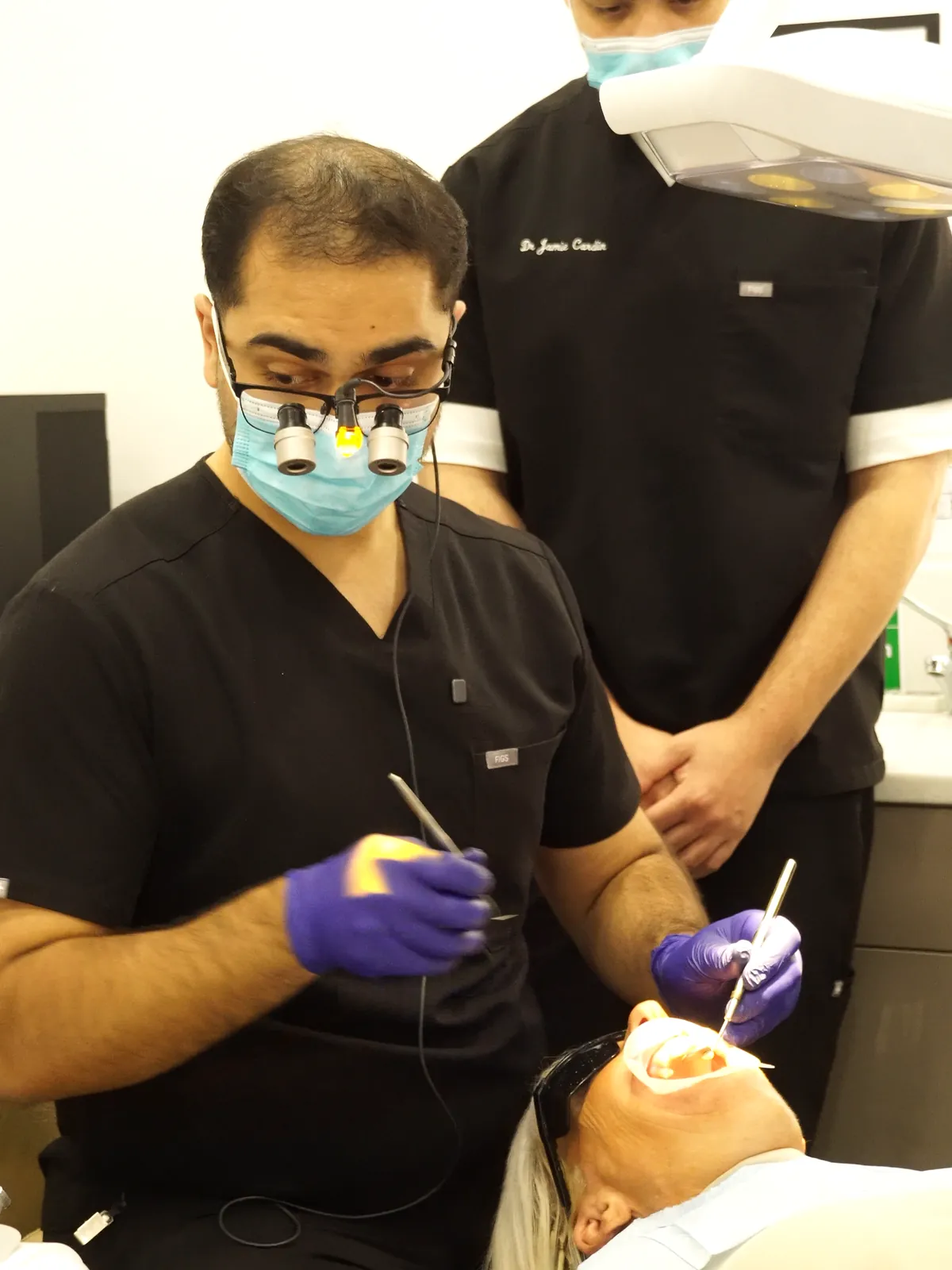 Dentist wearing loupes building up composite on June's front teeth, with a colleague observing